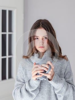 Vertical portrait of beautiful tween girl in warm grey pullover holding cup of tea looking at camera at home.