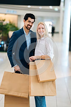 Vertical portrait of beautiful young couple holding shopping paper bags with purchases and looking at camera, standing