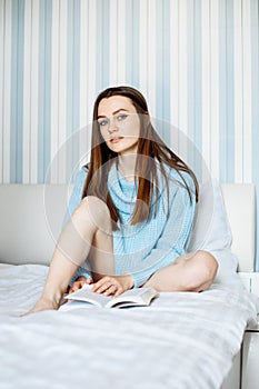 Vertical portrait of blue-eyed, thoughtful young woman in loose blue sweater sitting on bed with book in her hands.