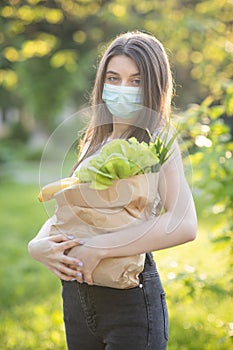 Vertical Portrait of Caucasian young pretty woman delivery worker in medical mask on the park carrying packet with fresh