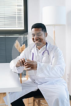 Vertical portrait of cheerful African-American male doctor wearing white uniform holding syringe in hand sitting at