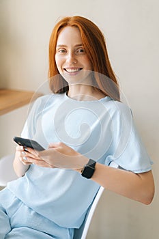 Vertical portrait of cheerful young woman in casual clothing holding mobile phone sitting at desk by window in cozy