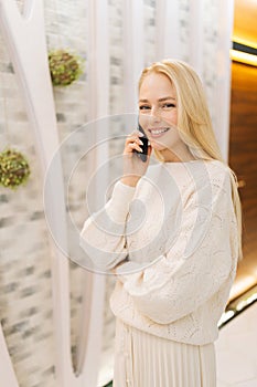 Vertical portrait of cheerful young woman talking on smartphone standing in hotel lounge, looking at camera. Cute blonde