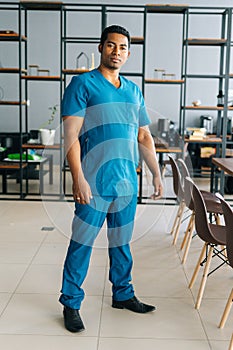 Vertical portrait of confident African American male doctor wearing blue surgeon medical uniform standing in hospital