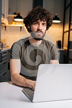 Vertical portrait of confident bearded young business man using on laptop sitting at table in kitchen room with modern