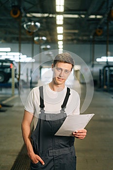 Vertical portrait of confident handsome young mechanic male wearing uniform holding clipboard standing in auto repair