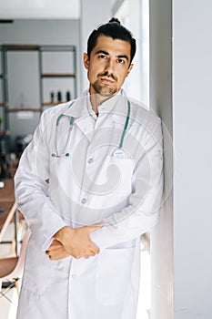 Vertical portrait of confident male doctor wearing white medical uniform standing with stethoscope in hospital office