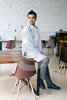 Vertical portrait of confident male doctor in white medical uniform sitting with stethoscope at table in hospital office