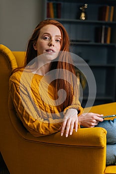 Vertical portrait of confident redhead woman using mobile phone sitting in yellow armchair looking at camera.