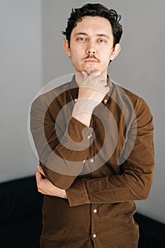 Vertical portrait of confident young man with little moustache holding hand on chin and thoughtful looking at camera