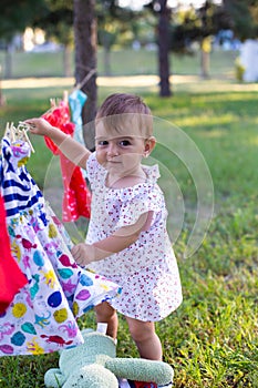 Vertical portrait of a cute baby girl in a summer dress against the background of summer nature and clothes that are dried in the