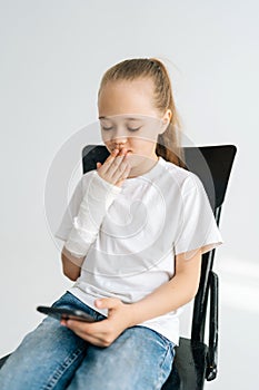 Vertical portrait of cute blonde little girl with broken arm wrapped in white plaster bandage looking at screen of