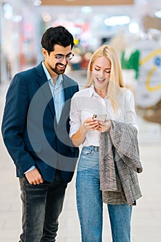 Vertical portrait of focused happy young couple using mobile phone standing in hall of shopping mall.