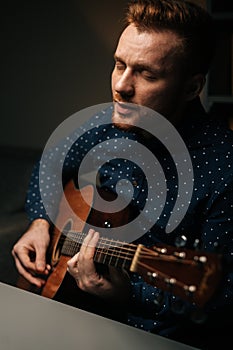 Vertical portrait of guitarist singer male playing acoustic guitar and singing song in dark living room at home studio.