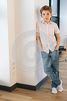 Vertical portrait of handsome child boy model leaning against wall keeping hands in jeans pockets and looking at camera