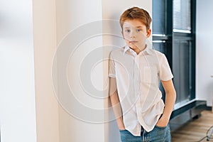 Vertical portrait of handsome child boy model leaning against wall keeping hands in jeans pockets and looking at camera