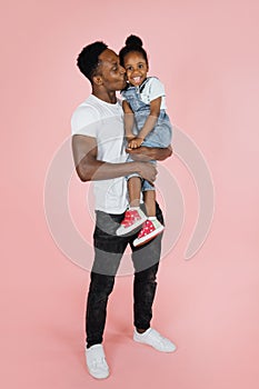 Vertical portrait of happy afro daddy embracing with his cute baby daughter, pink studio background.