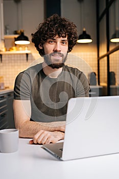 Vertical portrait of happy bearded young business man using typing on laptop sitting at table in kitchen room with