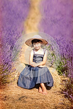 Vertical portrait of happy cute little girl wearing blue dress and white hat and sitting in lavender field with violet flowers