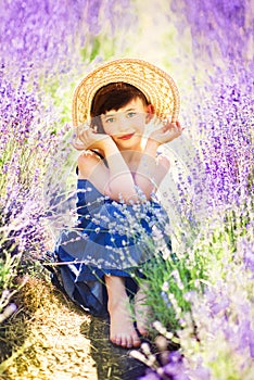 Vertical portrait of happy cute little girl wearing blue dress and white hat and sitting in lavender field with violet flowers