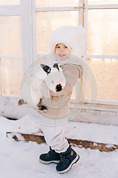 Vertical portrait of happy little child boy in warm clothes hugging white funny rabbit standing by farm house on sunny