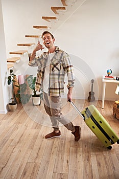 Vertical portrait of happy young man going on holiday, shows mobile phone call hand gesture, walking with suitcase