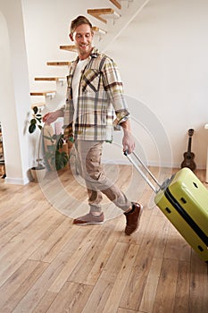 Vertical portrait of happy young man with suitcase, walking indoors, leaving a flat with luggage, going on business trip