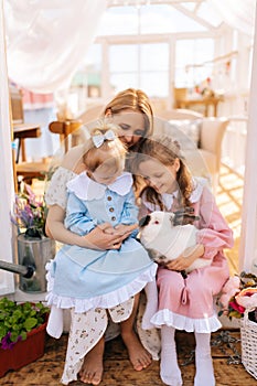 Vertical portrait of happy young mom and two adorable daughters in dress hugging rabbit in summer gazebo house on sunny