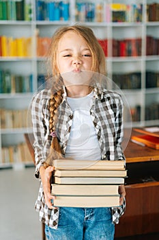 Vertical portrait of joyful elementary child school girl holding stack of books in library at school, looking at camera.