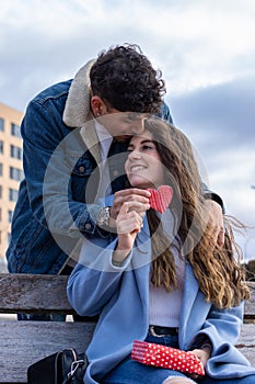 Vertical portrait of latin man hugging and kissing his girlfriend from behind giving her a surprise with a gift and a heart.