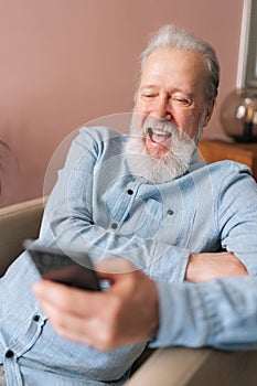 Vertical portrait of laughing mature male holding smartphone using mobile online app, cheerful looking to screen sitting