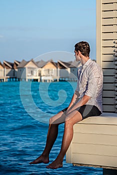 Vertical portrait of man wearing shirt sitting near water villas at the tropical beach at island luxury resort