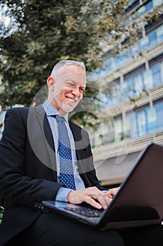 Vertical portrait of a mature businessman typing on a laptop outside. White collar worker man using a computer to work
