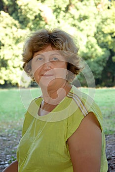 Vertical portrait of mature confident lady in green blouse looking at camera and smiling in the park, blurred background