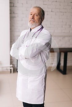 Vertical portrait of mature man doctor in white lab coat and stethoscope posing with crossed arms