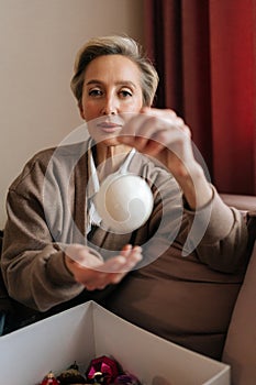 Vertical portrait of middle-aged woman holding Christmas ball, looking at camera with happy expression. Elegant female