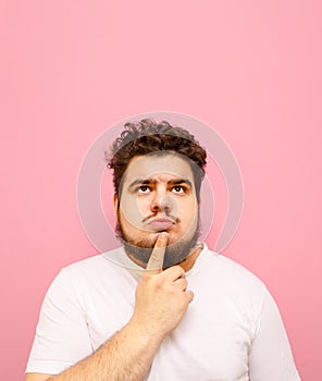 Vertical portrait of pensive young man with beard and overweight on pink background,looking up at copy space and thinking.Pensive