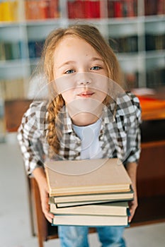 Vertical portrait of playful elementary child school girl holding stack of books in library at school, looking at camera