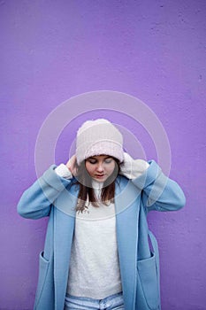 Vertical portrait of pleasent Hipster girl wearing warm casualblue coat and woolen hat on a lilac wall background.