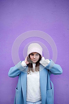 Vertical portrait of pleasent Hipster girl wearing warm casualblue coat and woolen hat on a lilac wall background. photo