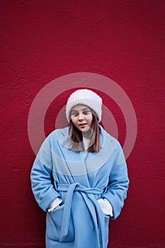 Vertical portrait of pleasent Hipster girl wearing warm casualblue coat and woolen hat on a vinous wall background.