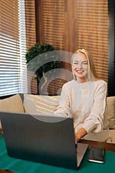 Vertical portrait of positive blonde young businesswoman sitting on sofa in cafe, working on laptop computer and smiling