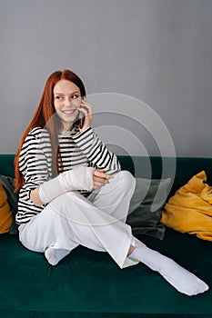 Vertical portrait of positive young woman with broken arm wrapped in gypsum bandage talking on smartphone with friends
