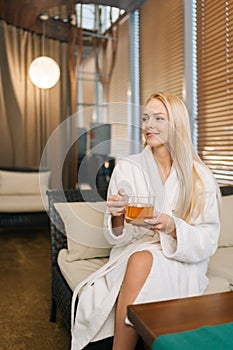 Vertical portrait of pretty blonde young woman in white bathrobe holding cup with fresh herbal tea smiling looking away