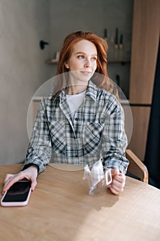 Vertical portrait of pretty young woman holding glasses mug with drip coffee sitting at kitchen table in morning