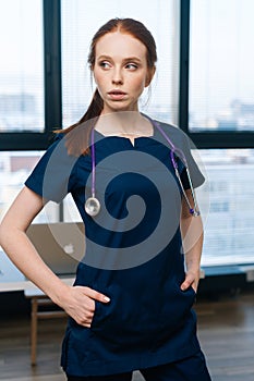 Vertical portrait of redhead serious female practitioner in medical uniform looking away standing on background of