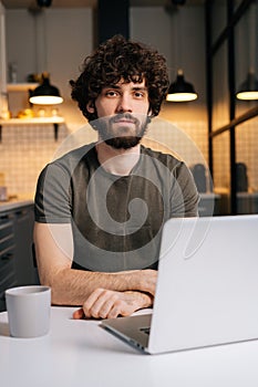 Vertical portrait of confident bearded young business man using on laptop sitting at table in kitchen room with modern