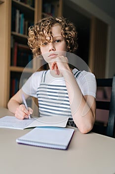 Vertical portrait of serious curly little school girl studying at home, doing homework, holding pen, writing notes
