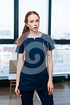 Vertical portrait of serious redhead female doctor wearing medical dark uniform looking at camera standing on background