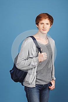 Vertical portrait of serious young red-headed male student in casual gray outfit with black backpack, holding hand in